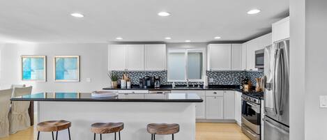 A modern kitchen with white cabinets, a black countertop, and stainless steel appliances. There are three wooden stools at the island, and a large window provides natural light.