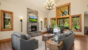 A spacious living room with a stone fireplace, two grey sofas, a wooden coffee table, and large windows offering a view of the outdoors. - Must See Suncadia Retreat! 3 Masters * Hot Tub * Game Room * On the Course! (Cle Elum)