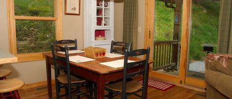 A dining area with a wooden table and chairs, a chandelier, and a view of greenery through a window.