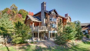 A large log cabin with a stone chimney, a covered porch, and a stone wall. - Ski-In/Ski-Out Creekside Condo with Private Hot Tub at Boyne Mountain (Boyne City)