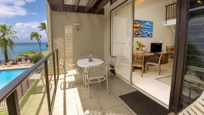Uma varanda com vista para a piscina, uma área de refeições com cadeiras e mesa, e uma porta de correr que dá para um espaço de estar com televisão e plantas.