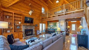 A spacious log cabin with a fireplace, a large sofa, a dining area, and a balcony. - Chipmunk Chase in Gated Smoky Mountain Retreat Community (MAGGIE VALLEY)
