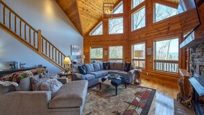 A spacious living room with a high wooden ceiling, large windows, a staircase, and a fireplace. - Chipmunk Chase in Gated Smoky Mountain Retreat Community (MAGGIE VALLEY)