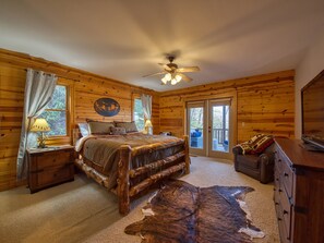 Un dormitorio tipo cabaña de madera con cama, cabecera de madera, ventilador de techo, ventana grande y vistas al exterior.