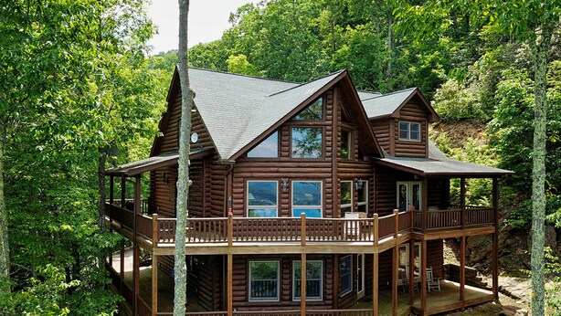 A log cabin with a balcony and multiple windows, surrounded by dense forest. - Chipmunk Chase in Gated Smoky Mountain Retreat Community (MAGGIE VALLEY)