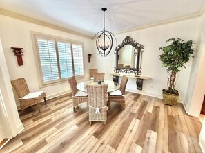 Une salle Ă manger avec des chaises en osier, une table ronde en verre, un lustre, un grand miroir et une plante en pot.