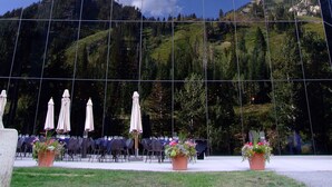 A modern building with a glass facade reflecting the sky and clouds. In front, there are outdoor tables with umbrellas and potted plants. - Prime Ski Week at Snowbird's Cliff Club Sleeps 6 in 1bedroom (Sandy)