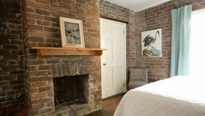 Ein Zimmer mit Kamin aus Ziegelsteinen, einer weißen Tür, einem gerahmten Bild über dem Kamin, einem Sessel und einem Bett mit weißer Bettdecke.