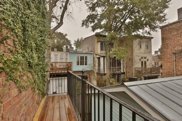 A narrow wooden deck with a black railing, surrounded by brick walls and various buildings. - Beautiful 1800s Carriage Home, Renovated w/ Modern Décor by Lucky Savannah (Savannah)