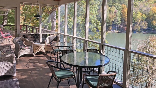 A covered outdoor seating area with wicker furniture and a glass table, overlooking a lake and forested area. - Lakefront Rumbling Bald Retreat, HotTub, Kayaks, SUP's, Floating Dock (Lake Lure)