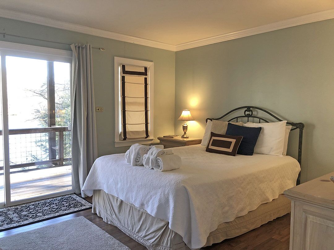 A neatly made bed with a white comforter and pillows, a bedside lamp, and a window with curtains.