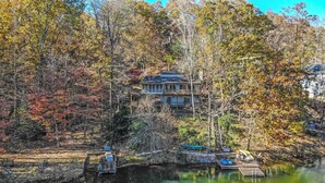 A two-story house surrounded by autumn foliage, with a dock and boats in the foreground. - Lakefront Rumbling Bald Retreat, HotTub, Kayaks, SUP's, Floating Dock (Lake Lure)