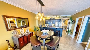 A dining area with a central table and benches, a kitchen in the background, and a mirror above the table. - Platinum, King Masters, Walk-In Showers, End-Unit Balcony, Free Beach Chairs! (Panama City Beach)