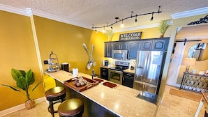 A kitchen with a central island, bar stools, stainless steel appliances, and a 'WELCOME' sign. - Platinum, King Masters, Walk-In Showers, End-Unit Balcony, Free Beach Chairs! (Panama City Beach)