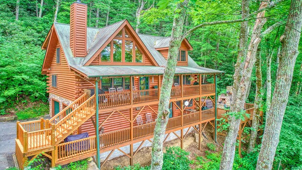 A log cabin with multiple decks and outdoor seating areas, surrounded by dense forest.
