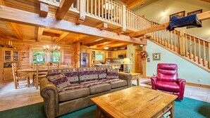A spacious cabin interior with a wooden staircase, a dining area, a living room with a sofa and a red leather chair, and a kitchen in the background.