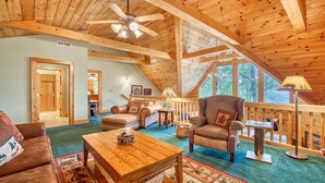 A cozy cabin interior with a high ceiling, wooden beams, a large window, and a furnished living area including a sofa, armchair, and coffee table.