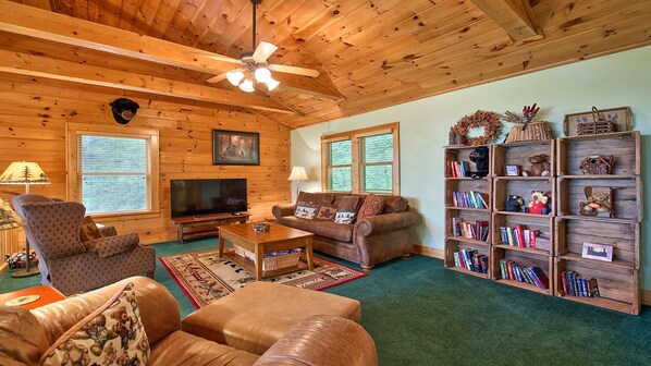 A cozy living room with wooden walls and ceiling, a television, leather sofas, a coffee table, and a bookshelf with books and decorative items.