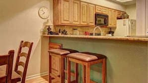 Una cocina con armarios de madera, un microondas, un refrigerador, un reloj y dos taburetes de bar.