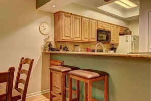 Une cuisine avec des meubles de rangement en bois, un four à micro-ondes, un réfrigérateur, une horloge et deux tabourets de bar.