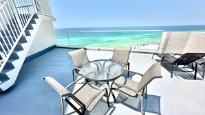A sunlit outdoor seating area with a glass table and wicker chairs, overlooking a beach and turquoise waters. - Fabulous gulf/beach views from the 14th floor! 4336BS2 (Miramar Beach)
