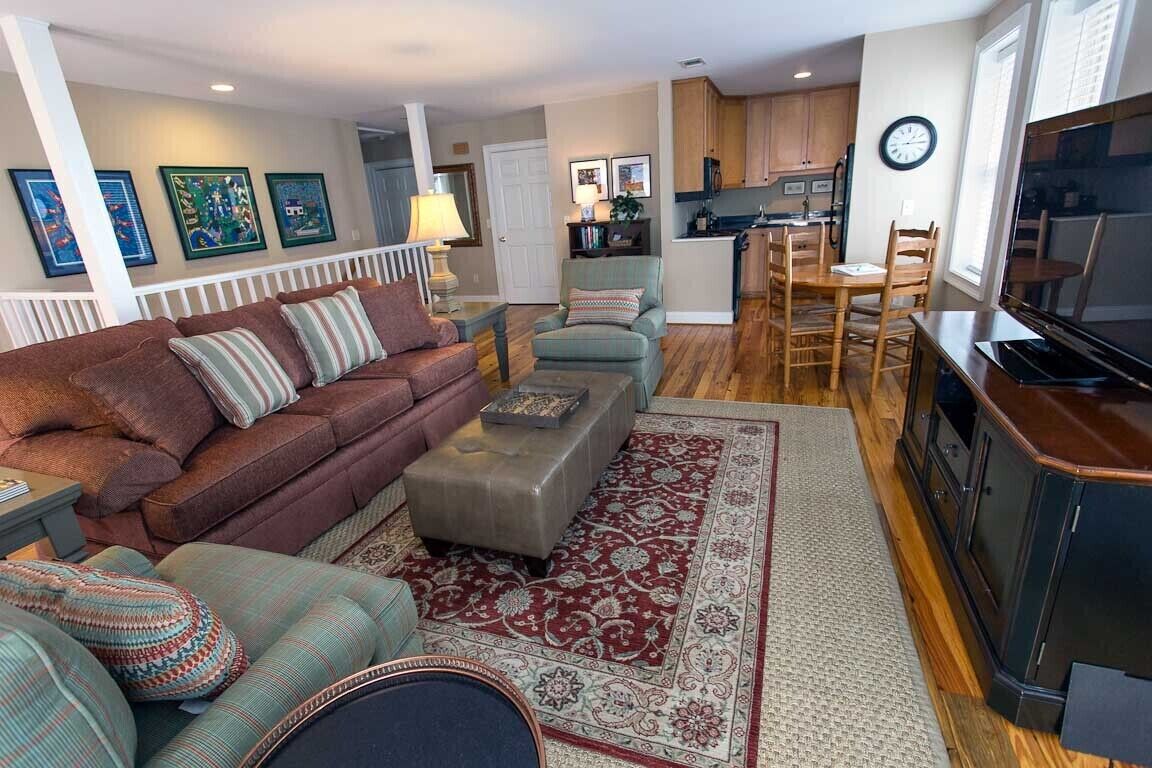 A living room with a sofa, ottoman, and chairs, a dining area with a table and chairs, a kitchen with cabinets and appliances, and a TV.