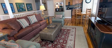 Een woonkamer met een bank, poef en stoelen, een eethoek met tafel en stoelen, een keuken met kasten en apparatuur, en een televisie.