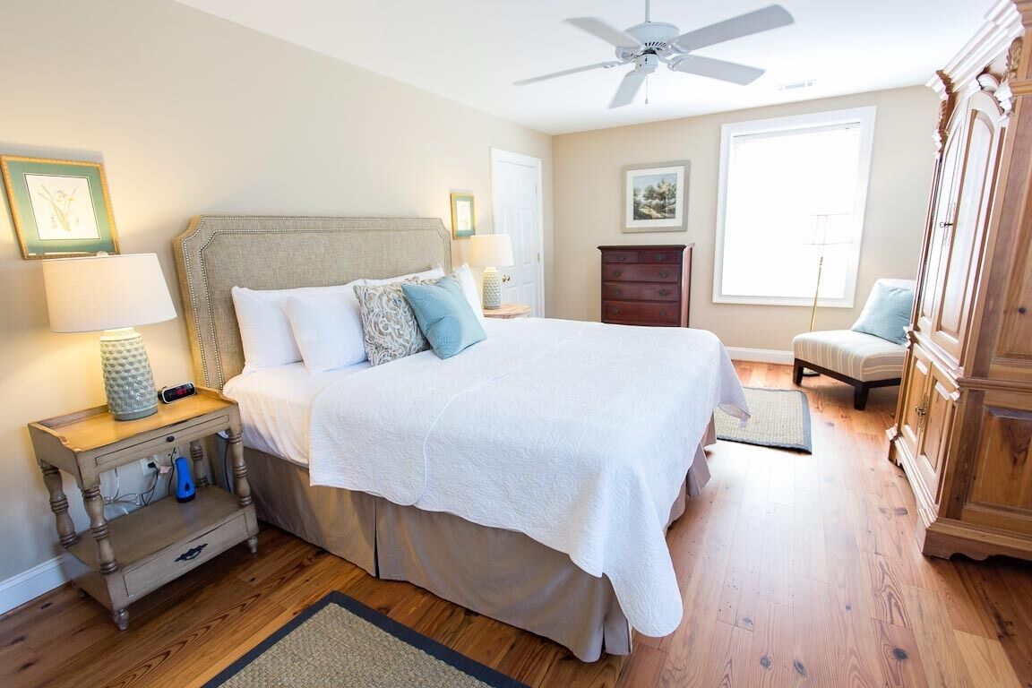 Une chambre à coucher avec un grand lit, une table de chevet avec une lampe, une commode et un ventilateur de plafond.
