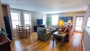 A living room with a sofa, armchair, dining table, and chairs. There is a television and a plant.