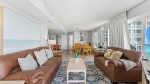 A spacious living room with two brown leather sofas, a wooden coffee table, and a patterned rug. There is a dining area with wooden chairs and a chandelier. Large windows provide a view of the sea. - Family fun with amazing amenities + Free Attraction Tickets! (Myrtle Beach)