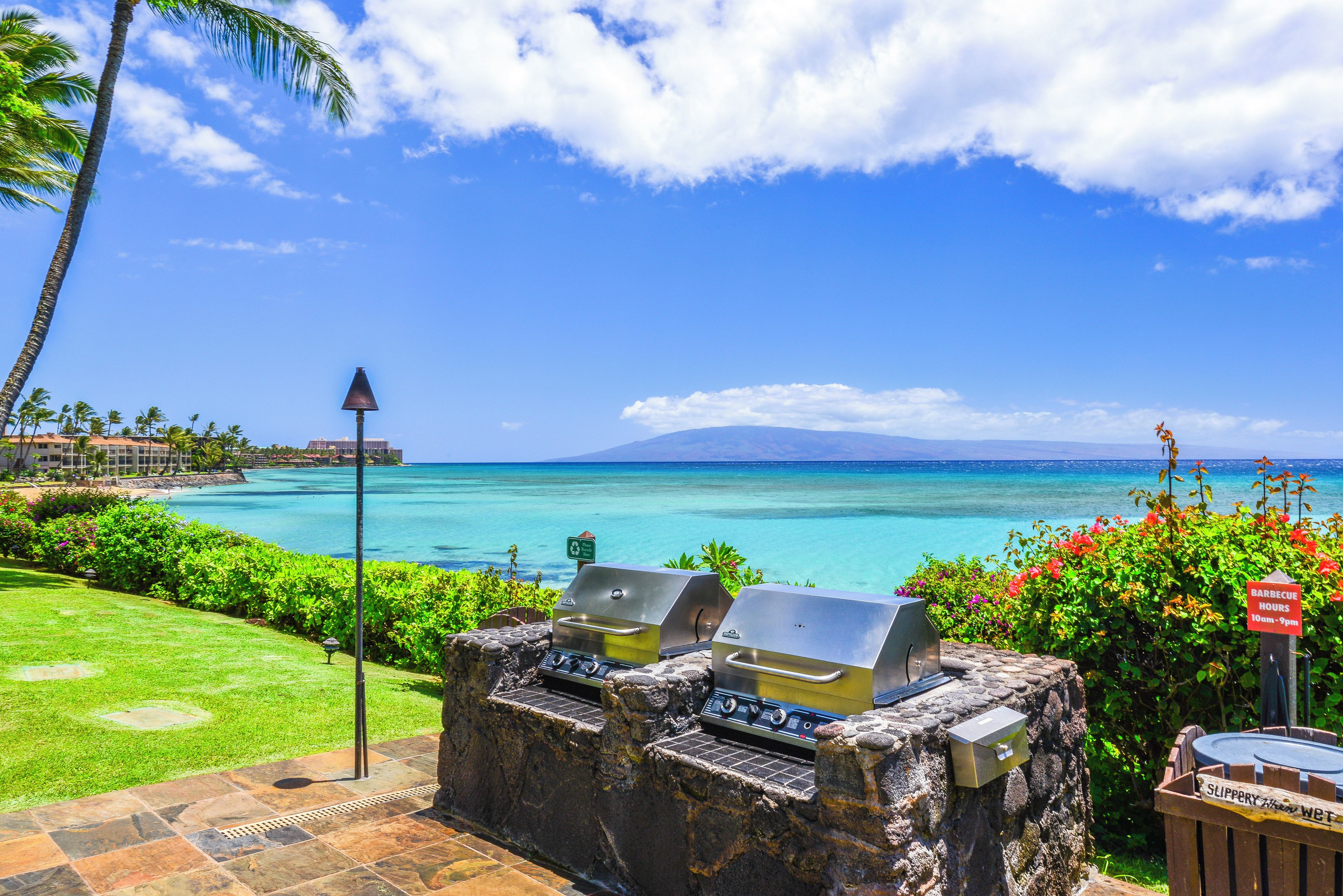 A stone barbecue area with two grills, a clear view of the ocean, and a tropical setting with palm trees and a sign indicating barbecue hours.