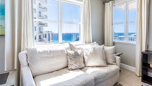 Una sala de estar moderna con un sofá beige, dos ventanas con cortinas y vistas al océano y edificios.
