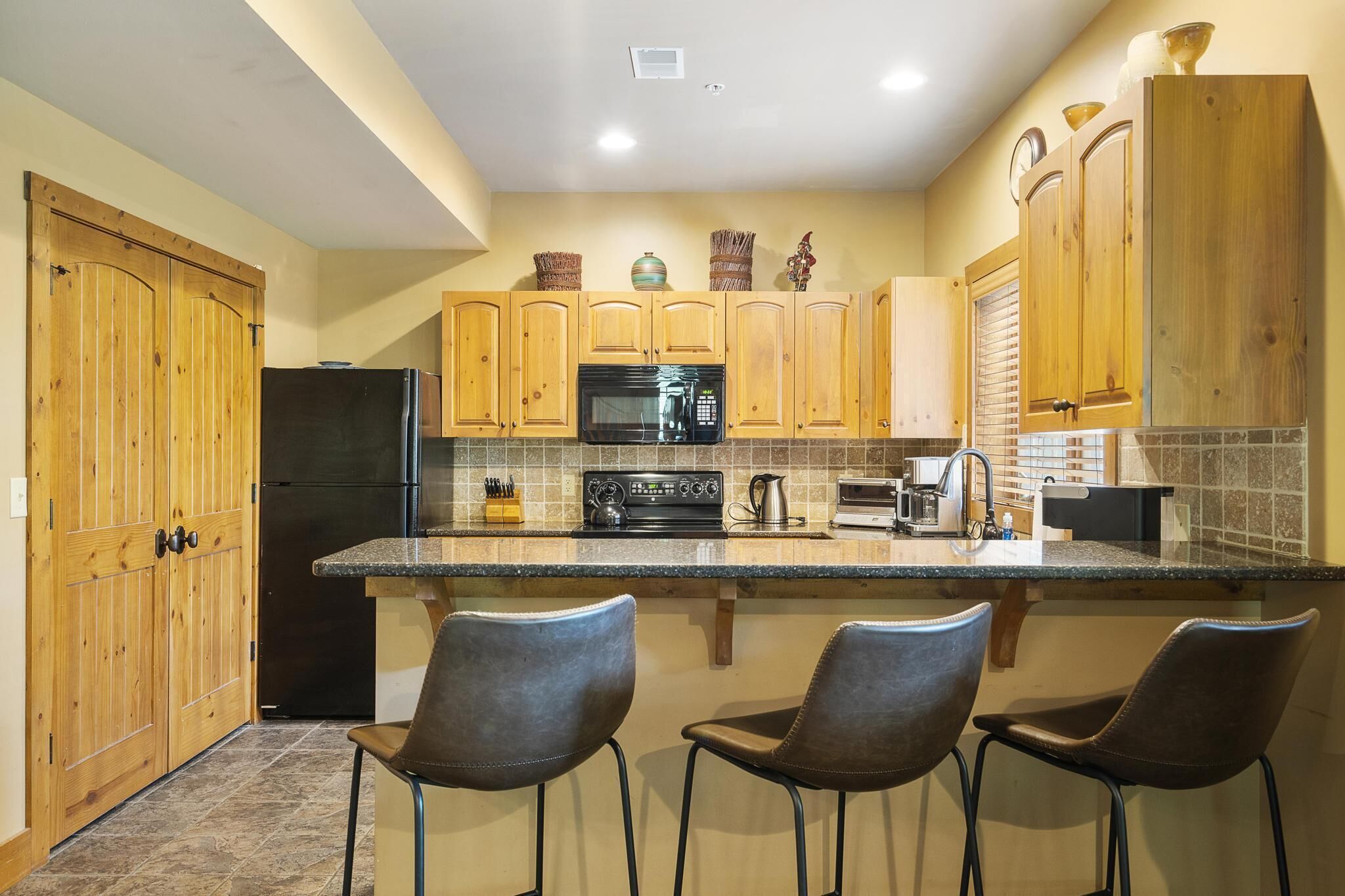 A kitchen with wooden cabinets, a black refrigerator, a microwave, a stove, and a coffee maker. There are two bar stools at the island.