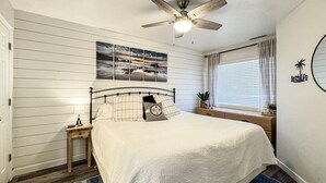 A bedroom with a bed, a nightstand, a ceiling fan, a window with curtains, and a wall-mounted artwork.