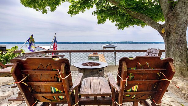 A wooden deck with two chairs and a table, overlooking a lake with a gazebo and a dock in the background. - Seneca Sunsets - Awesome Lakeside Getaway Near Geneva! (Geneva)