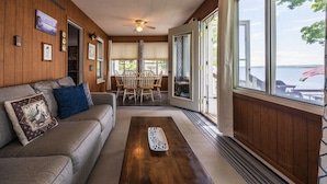 A cozy living room with a wooden floor, a gray sofa with decorative pillows, a wooden coffee table, and a dining area with a table and chairs. - Seneca Sunsets - Awesome Lakeside Getaway Near Geneva! (Geneva)