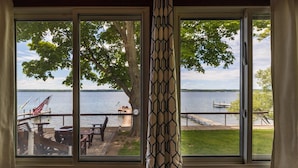 A view through a sliding glass door of a deck with outdoor furniture, a tree, and a lake. - Seneca Sunsets - Awesome Lakeside Getaway Near Geneva! (Geneva)