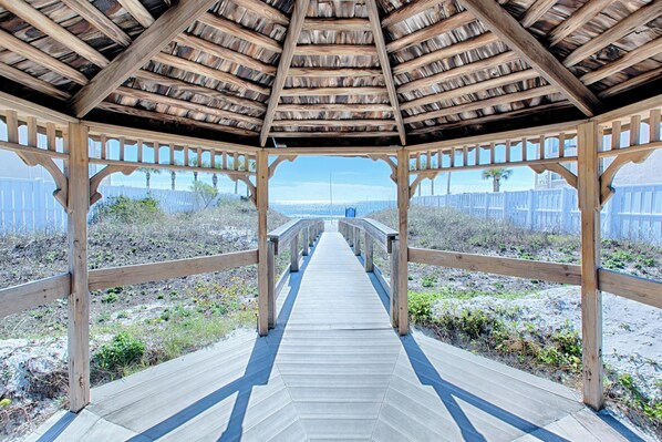A wooden deck leading to a covered area with a view of a sandy beach and a distant building.