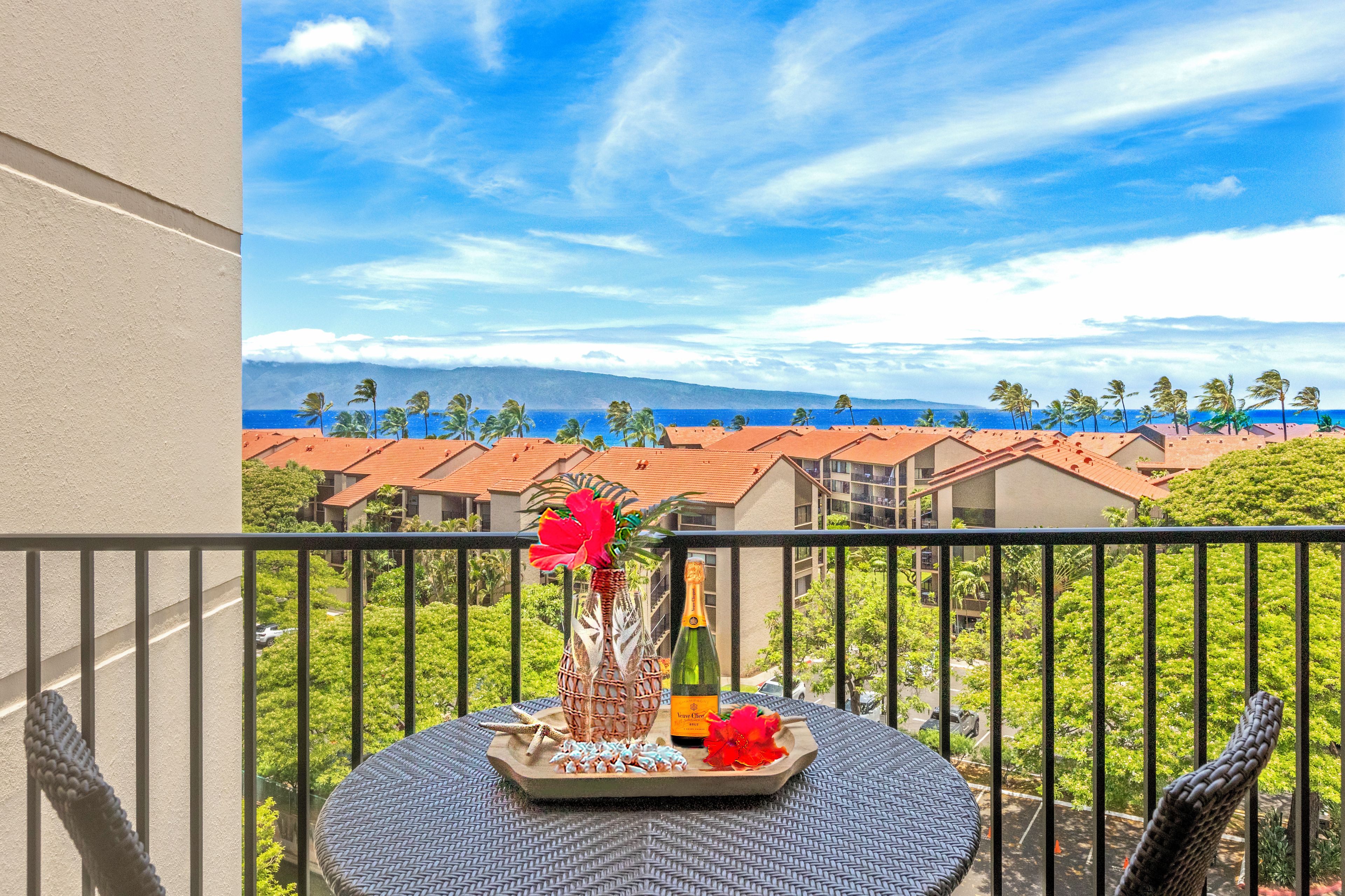 Ein Balkon mit Tisch für zwei Personen, ausgestattet mit einer Flasche, einer Blumenvase und einer Platte mit Snacks. Blick auf einen Strand und Palmen.