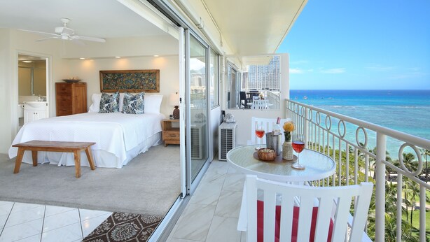 A balcony with a table and chairs, a bed with a view of the ocean, and a painting on the wall.