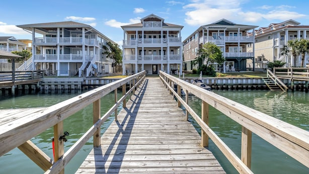 Un muelle de madera que conduce a una propiedad frente al mar, con varios balcones y un cielo despejado.