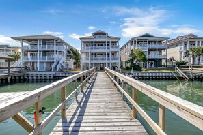 A wooden pier leading to a waterfront property with multiple balconies and a clear sky.