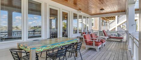 A covered outdoor area with a wooden deck, a long table with chairs, and a seating area with red and white chairs.