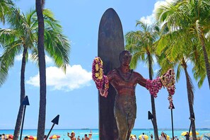 A statue of a man with a surfboard, surrounded by palm trees and beach umbrellas.