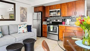 A compact kitchen with wooden cabinets, a stainless steel refrigerator, a microwave, and an oven. There is a gray sofa with colorful cushions, a wicker chair, and a glass table with a vase of flowers. - Ocean View Lanai and an Easy 5–10-min walk to beach! FREE Wi-Fi, Full Kitchen (Honolulu)