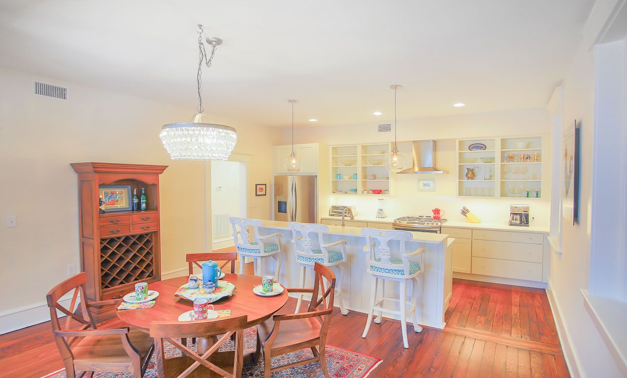 A dining area with a wooden table and chairs, a chandelier, and a kitchen in the background.