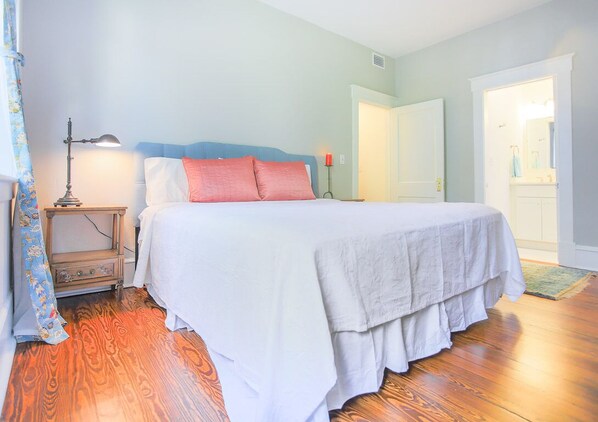 A bedroom with a bed, a nightstand, a lamp, and a door leading to a bathroom. - Beautiful, Centrally Located, Design District Apartment by Lucky Savannah (Savannah)