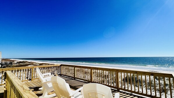 A wooden deck with white plastic chairs overlooking a beach with a clear blue sky.