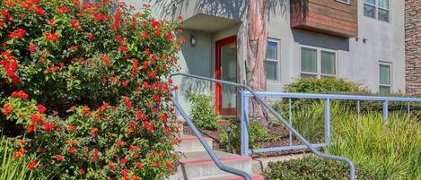Un edificio con puerta roja, una escalera con barandilla azul y un jardín con flores rojas.