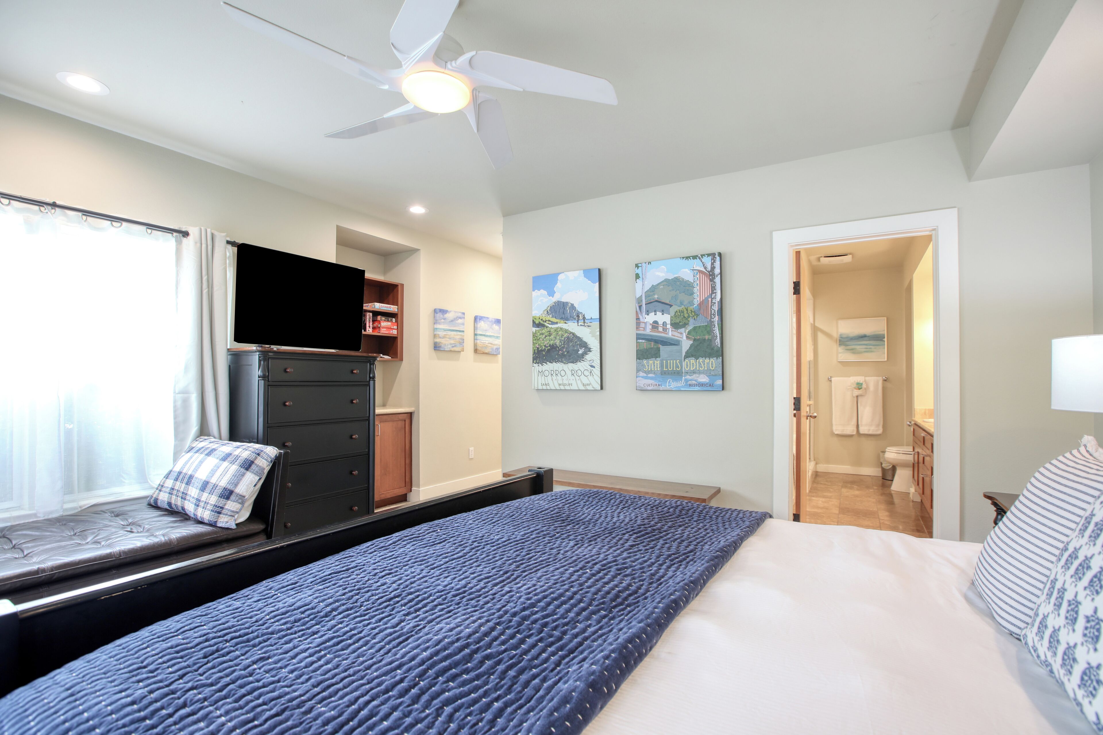 A bedroom with a bed, a ceiling fan, a window with curtains, a dresser, and artwork on the wall.